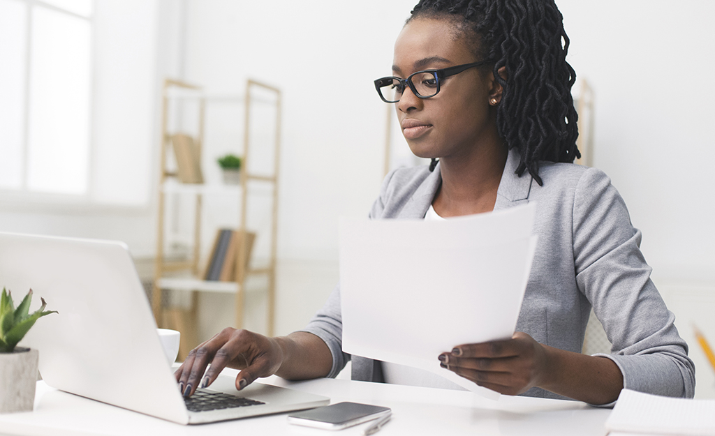 Mulher negra, cabelos longos, blusa branca manga comprida, usando óculos, com papel na mão e um notebook em frente, posto em cima de uma mesa branca.