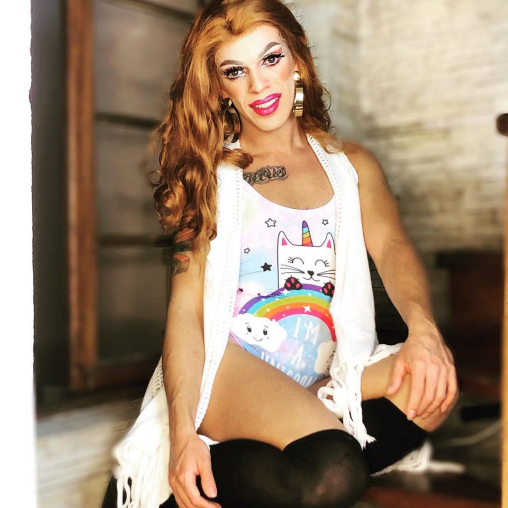 Rita DLibra vestindo coturno preto, maiô branco com estampa, cardigan longo branco, maquiagem bem marcada no rosto. Sentada em uma mesa de madeira, fazendo pose para a câmera.